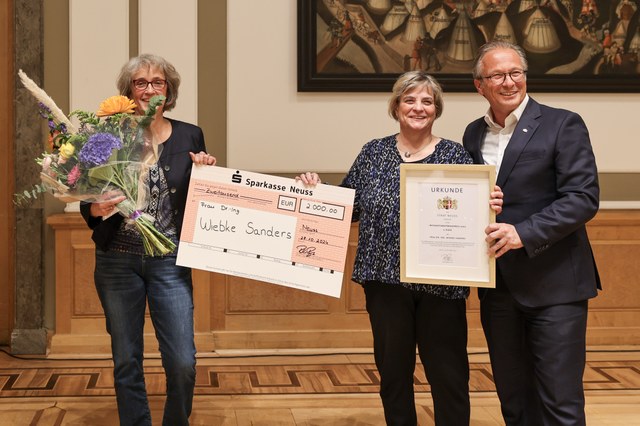 Die Preisträgerin hält einen Blumenstrauß in der Hand und zeigt gemeinsam mit dem Bürgermeister einen symbolischen Scheck sowie die Urkunde in die Kamera.