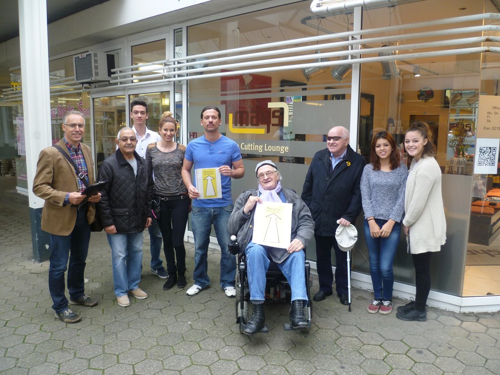 Personen auf dem Bild (von links): Harald Jansen (Projektleiter „Neuss barrierefrei“)  Inhaber Murat Alemdar (im blauen Shirt mit Signet) mit Familie und Team, Harry Aron -im Rollstuhl sitzend und Bernd Lynen mit dunkler Brille (beide Begehungsteam)