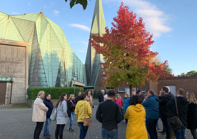 Eine größere Gruppe Menschen versammelt sich vor einer modernen Kirche und hört einer Person zu. Es ist ein sonniger Tag, im Hintergrund steht ein Baum dessen Blätter sich bereits zur Hälfte herbstlich rot gefärbt haben.