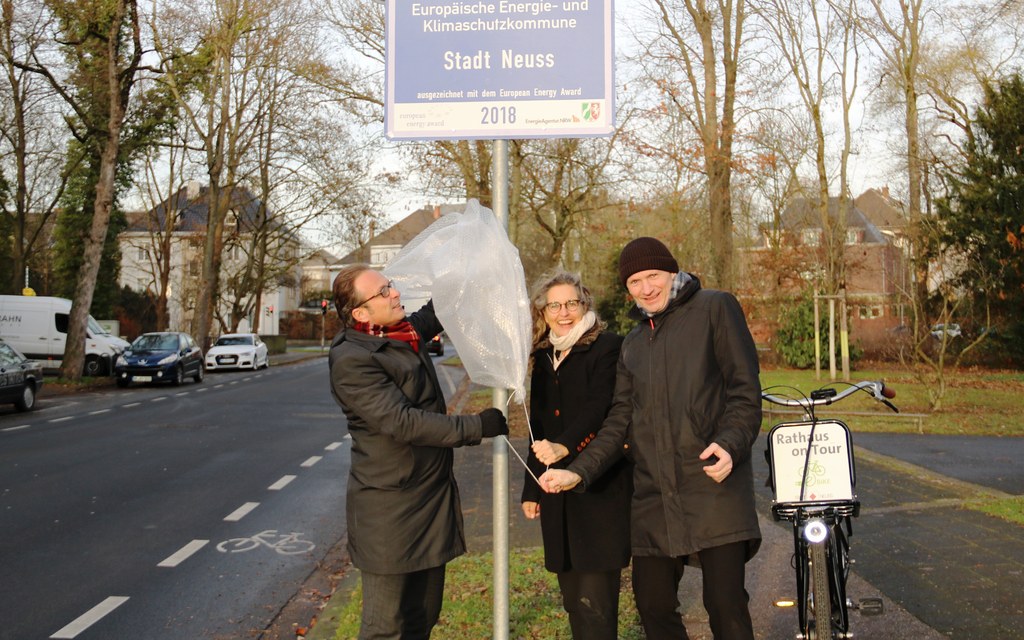 Bürgermeister Reiner Breuer (links), Umweltdezernent Dr. Matthias Welpmann und Henrike Mölleken, Leiterin des Amtes für Stadtgrün, Umwelt und Klima, enthüllen das Schild der EEA-Auszeichnung 2018 auf der Deutschen Straße.